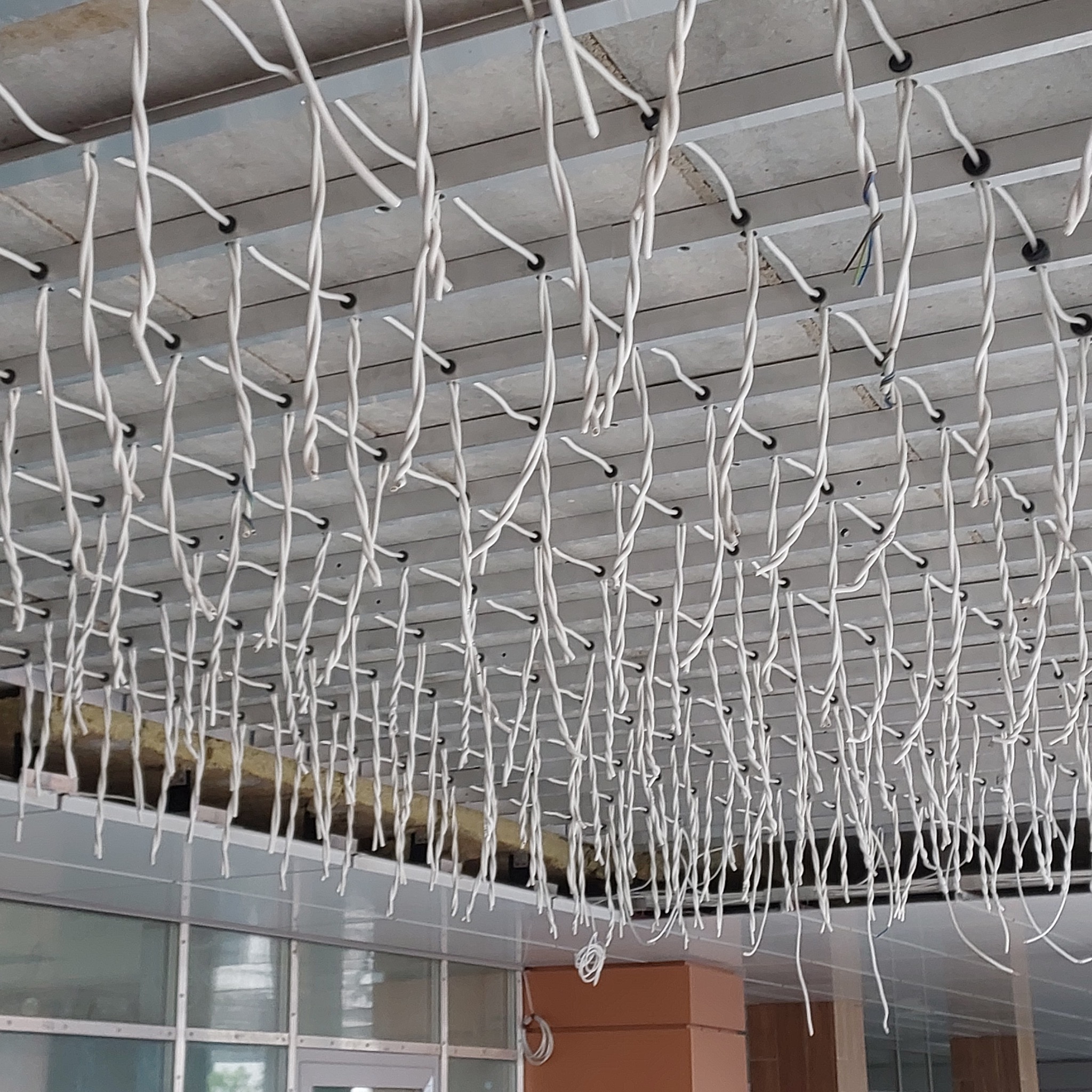 Zwisy kabli elektrycznych przymocowane do metalowej konstrukcji sufitu podwieszanego, widoczne fragmenty instalacji i okna.