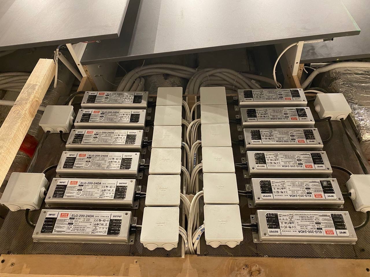 Rząd srebrnych zasilaczy LED (model ELG-200-24DA) przymocowanych do drewnianej konstrukcji pod sufitem, z białymi puszkami połączeniowymi i uporządkowanymi kablami.