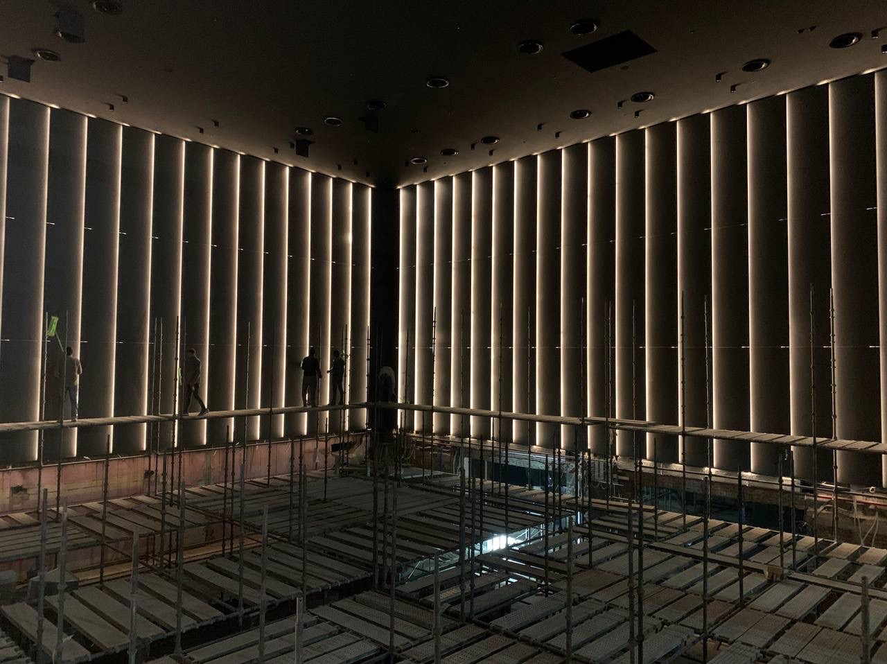 Montaż pionowego oświetlenia LED na ścianie w dużej sali, widoczne rusztowania i pracownicy w oddali.