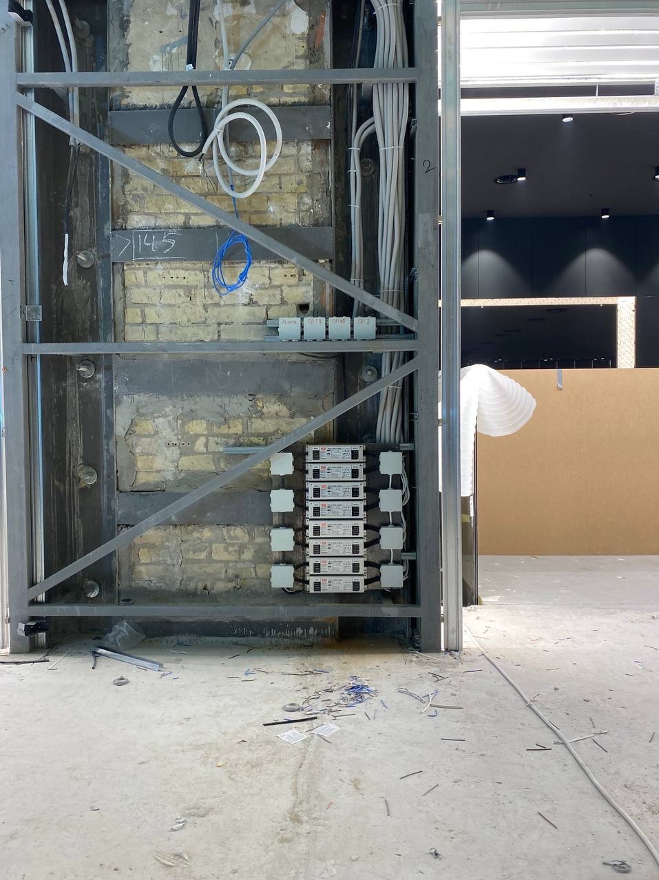 Surowy mur z cegły z metalową konstrukcją, widoczne kable elektryczne, transformatory i przewody, podłoże betonowe z resztkami izolacji i przewodów, wnętrze w trakcie prac wykończeniowych.