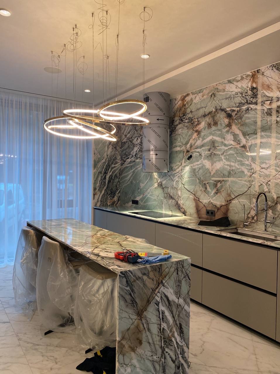 Luksusowa kuchnia z marmurowymi ścianami i wyspą, wiszące, okrągłe lampy LED, krzesła zabezpieczone folią, narzędzia na blacie, okap kuchenny zabezpieczony folią ochronną.