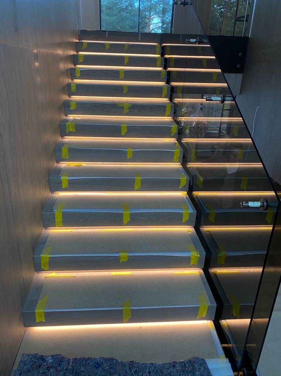 Nowoczesne schody drewniane z podświetleniem LED na każdym stopniu, zabezpieczone taśmą malarską, z szklaną balustradą, widok z dołu.