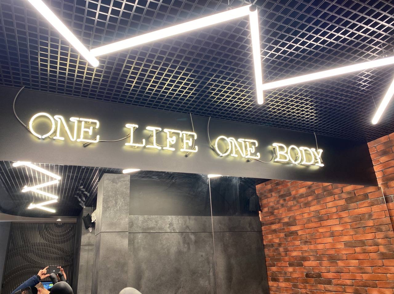 Neonowy napis 'ONE LIFE ONE BODY' pod sufitem z oświetleniem rastrowym w pomieszczeniu z ceglaną ścianą i szarą okładziną.