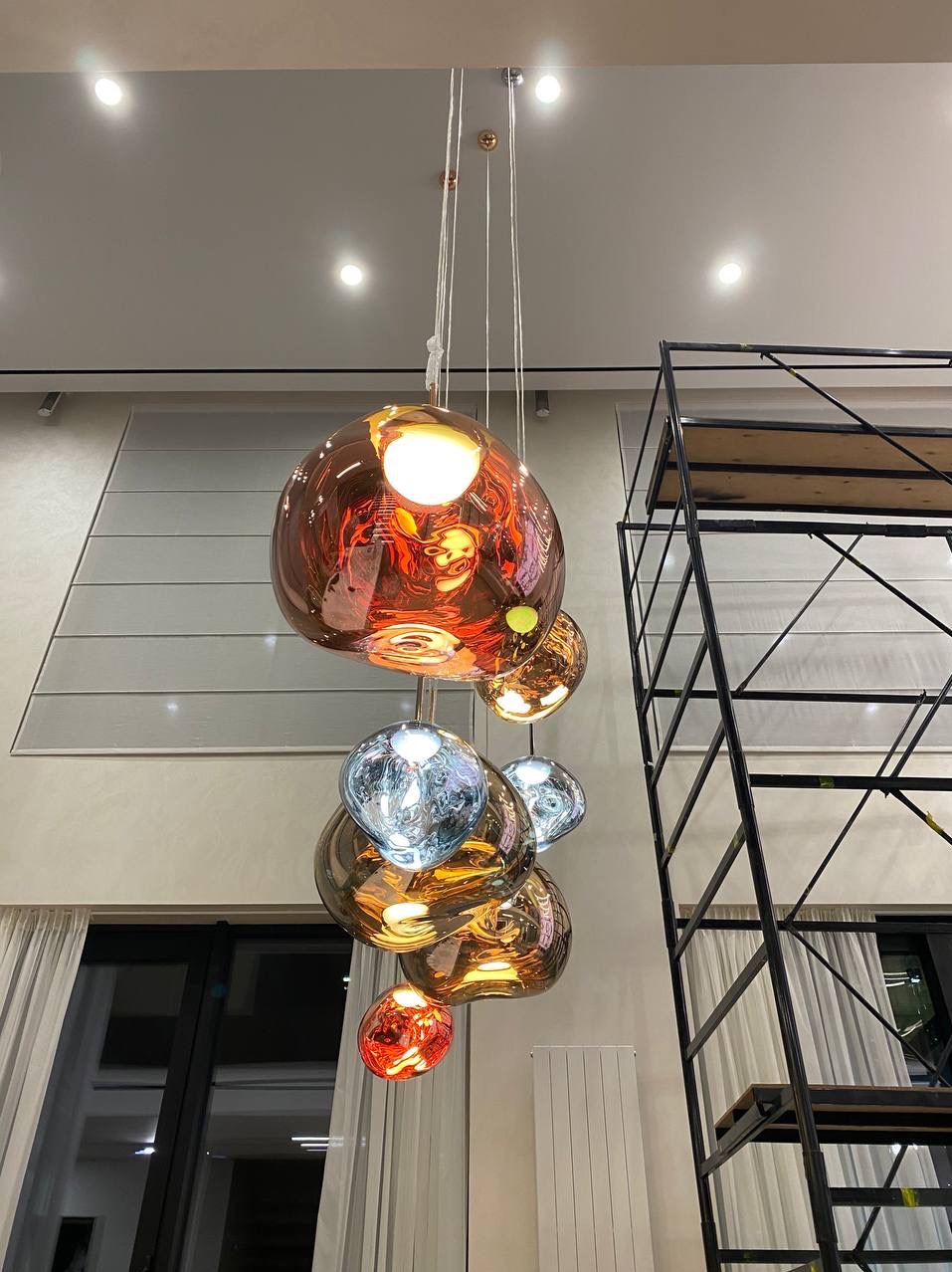 Kaskada szklanych, wielobarwnych kul tworzących designerską lampę wiszącą na tle jasnego wnętrza z wysokim sufitem i rusztowaniem malarskim.