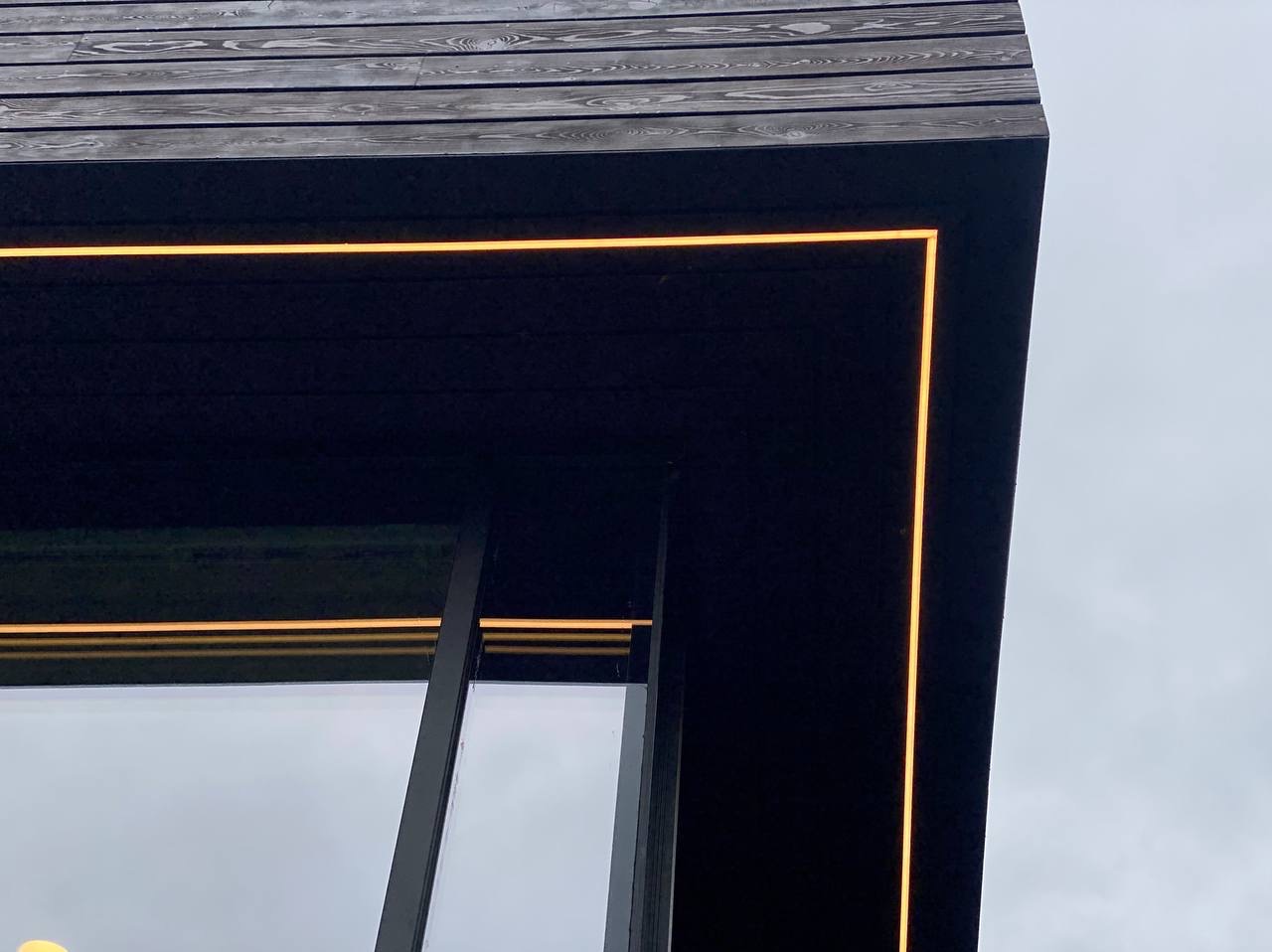 Detale elewacji budynku z ciemnego drewna z oknem i liniowym oświetleniem LED w ciepłej barwie, widok z dołu pod kątem.