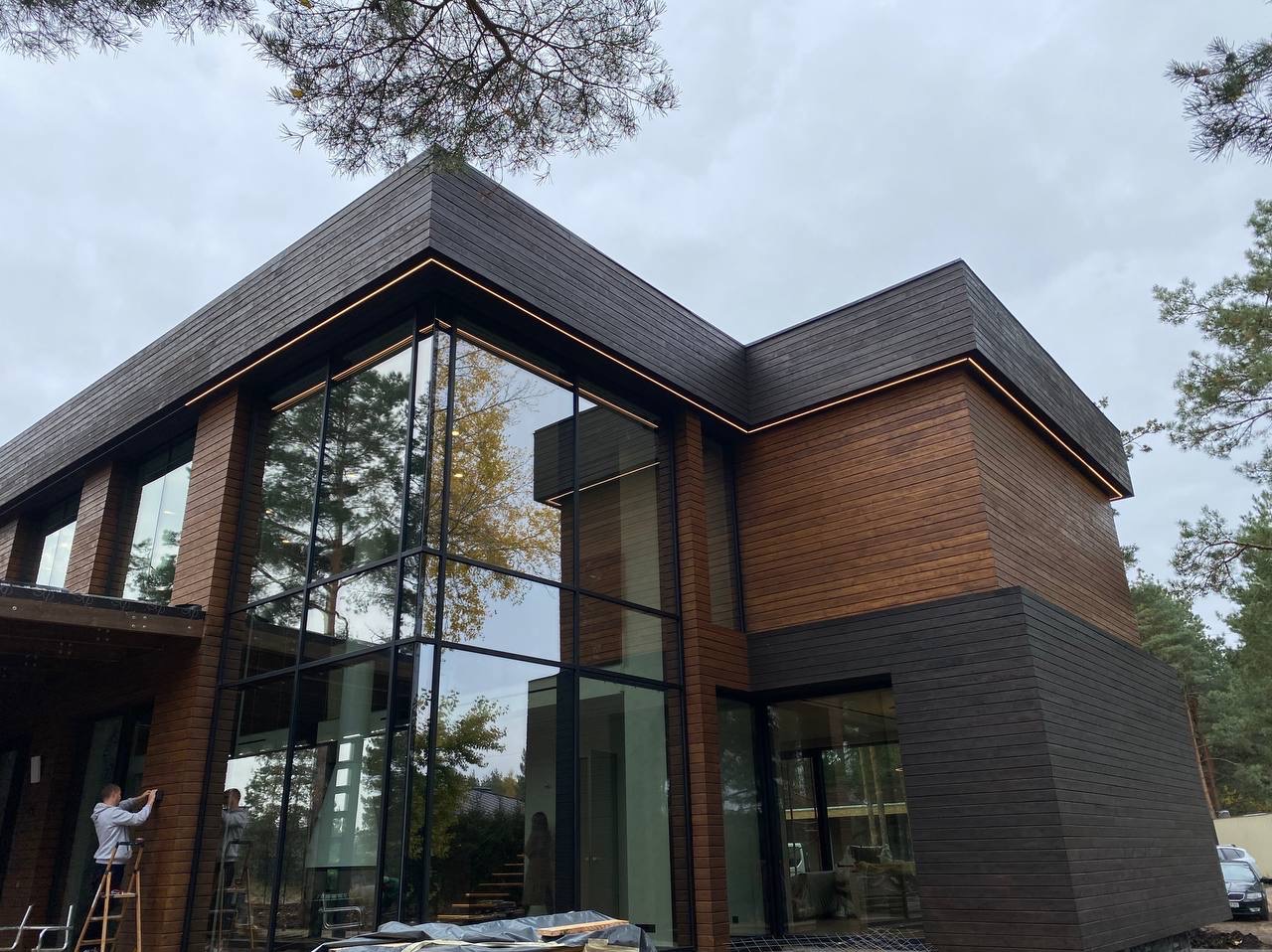 Nowoczesny dom z drewnianą elewacją i dużymi oknami, podświetlony ledami, pracownik na drabinie przy ścianie.