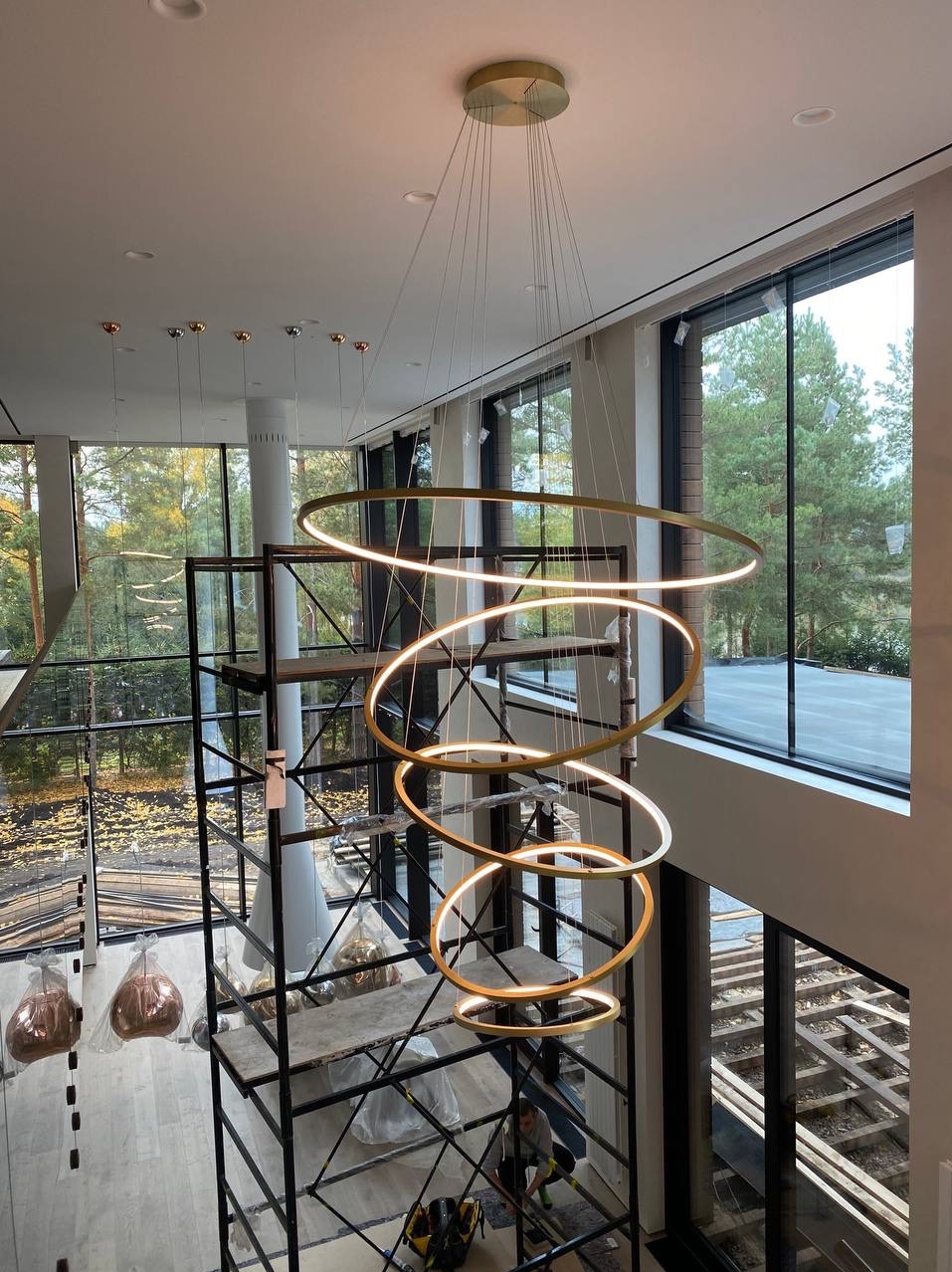 Wysoki hol w Zielonej Górze z nowoczesnym, spiralnym żyrandolem LED z metalowymi obręczami, widok z góry na rusztowanie i pracownika podczas montażu oświetlenia.