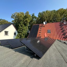 Energy Life Valentyn Shutko - Częściowo zamontowane czarne panele na płaskim dachu pokrytym papą, w tle dom z białymi ścianami i dachówką oraz dom z czerwoną dachówką i anteną satelitarną, błękitne niebo.