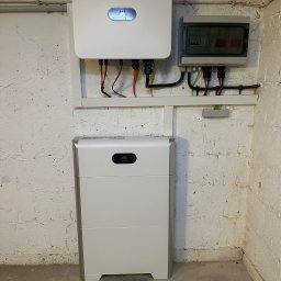 Energy Life Valentyn Shutko - Biały falownik i akumulator energii z logo Huawei zamontowane na ścianie z białej cegły, obok szara skrzynka z bezpiecznikami.