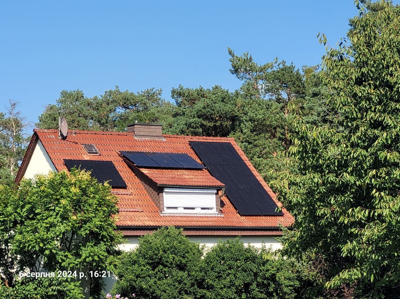 Częściowy widok dachu z pomarańczowej dachówki, na którym zamontowano czarne panele słoneczne, otoczonego zielenią drzew i krzewów, z widoczną anteną satelitarną.