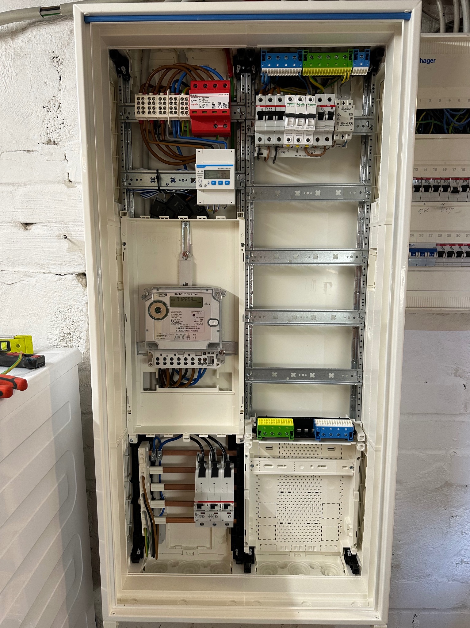 Otwarta skrzynka elektryczna z zamontowanym licznikiem energii, bezpiecznikami i okablowaniem, widok z bliska na białą ścianę z cegły w tle.