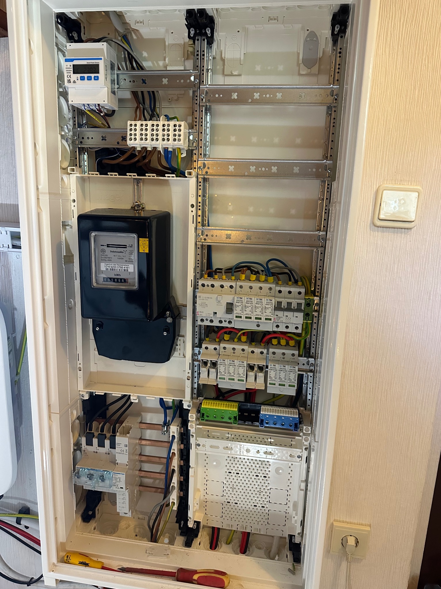 Otwarta skrzynka rozdzielcza z zainstalowanym licznikiem energii elektrycznej, bezpiecznikami i przewodami, widoczny śrubokręt na dnie obudowy.