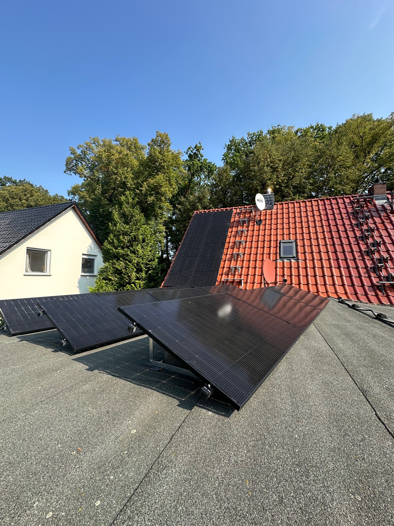 Częściowo zamontowane czarne panele na płaskim dachu pokrytym papą, w tle dom z białymi ścianami i dachówką oraz dom z czerwoną dachówką i anteną satelitarną, błękitne niebo.