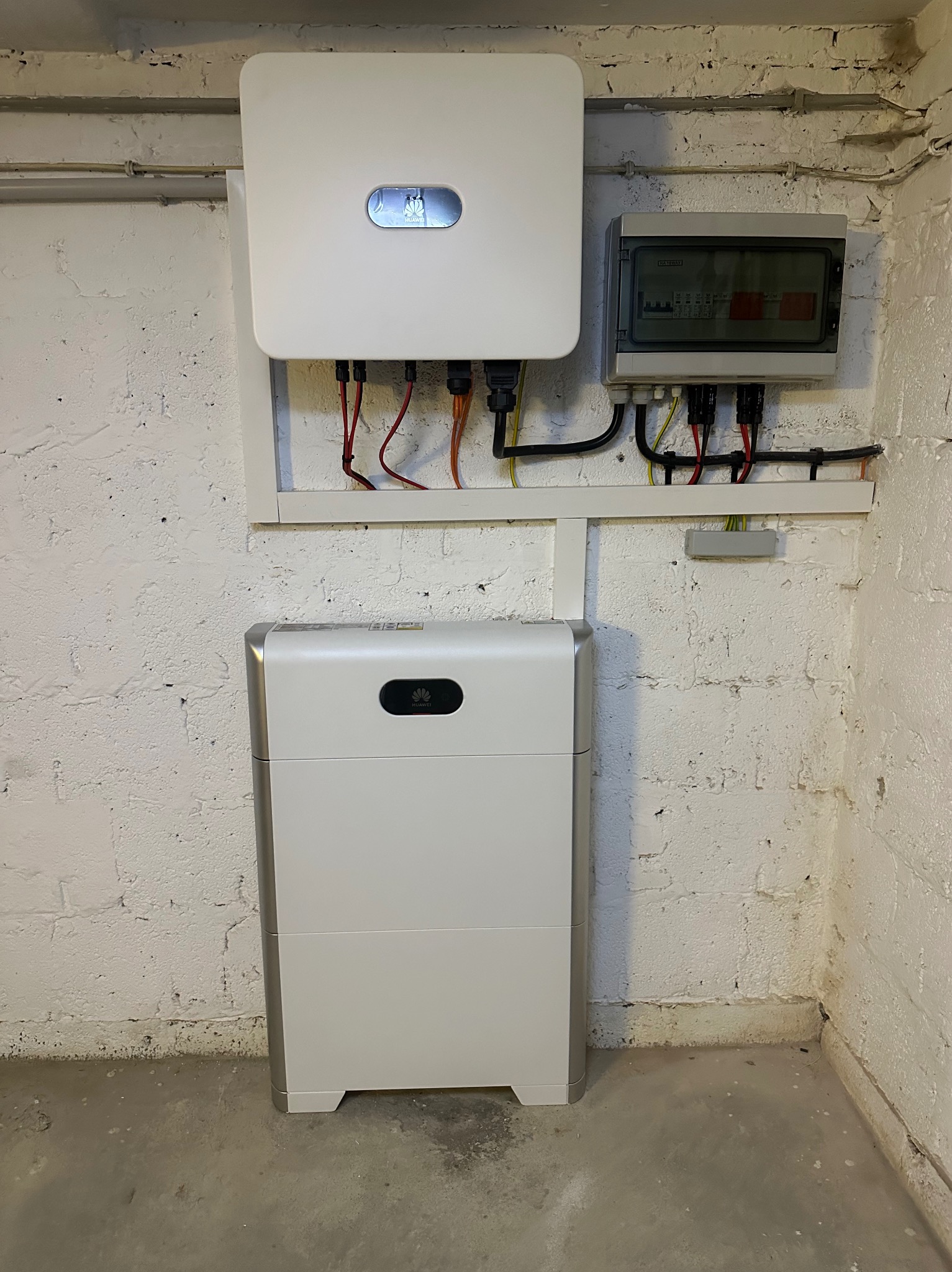 Biały falownik i akumulator energii z logo Huawei zamontowane na ścianie z białej cegły, obok szara skrzynka z bezpiecznikami.