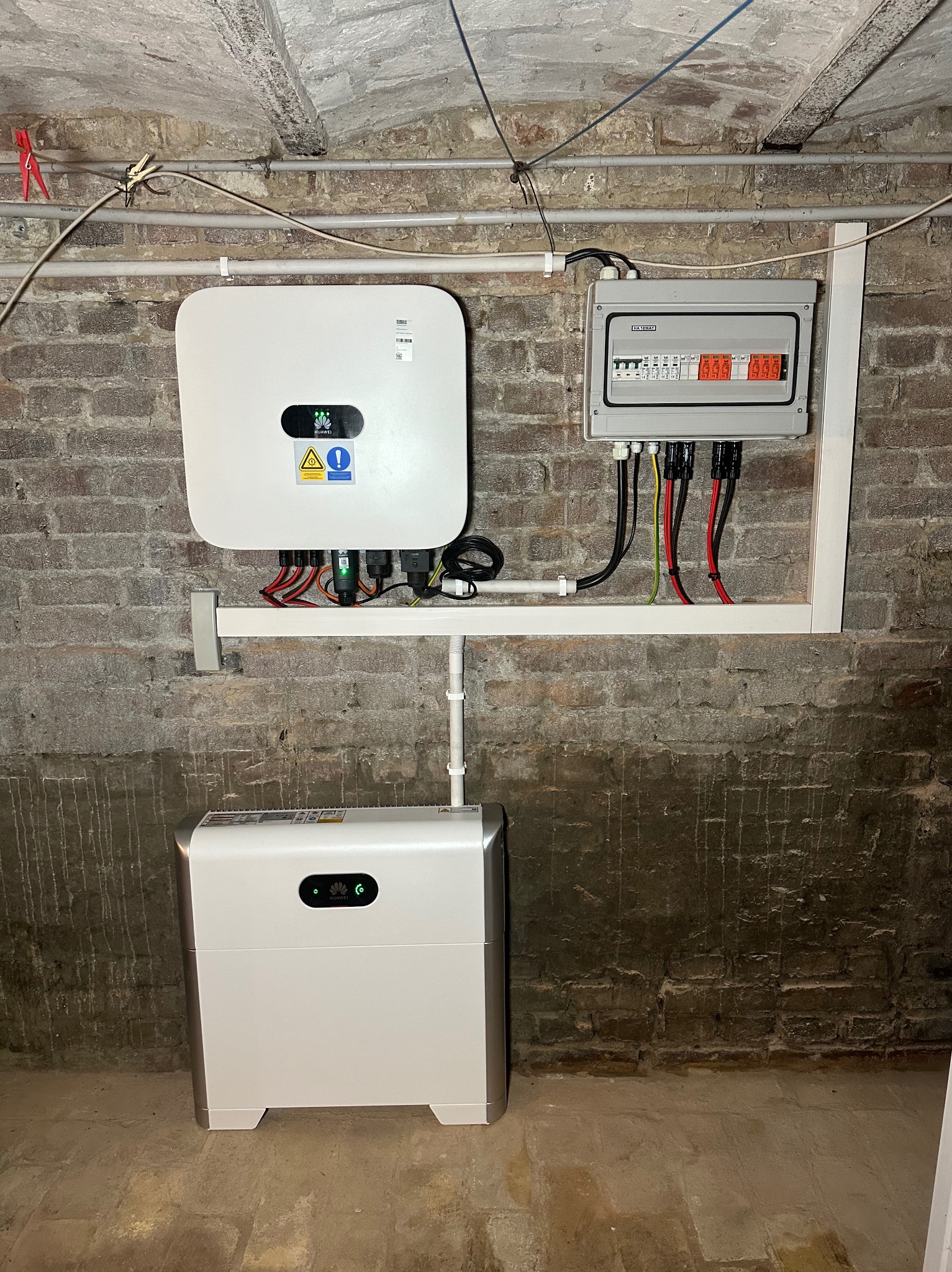 Kompletna instalacja fotowoltaiczna w piwnicy z białym inwerterem Huawei, akumulatorem i skrzynką bezpiecznikową na ceglanej ścianie, widoczne kable i rurki instalacyjne.