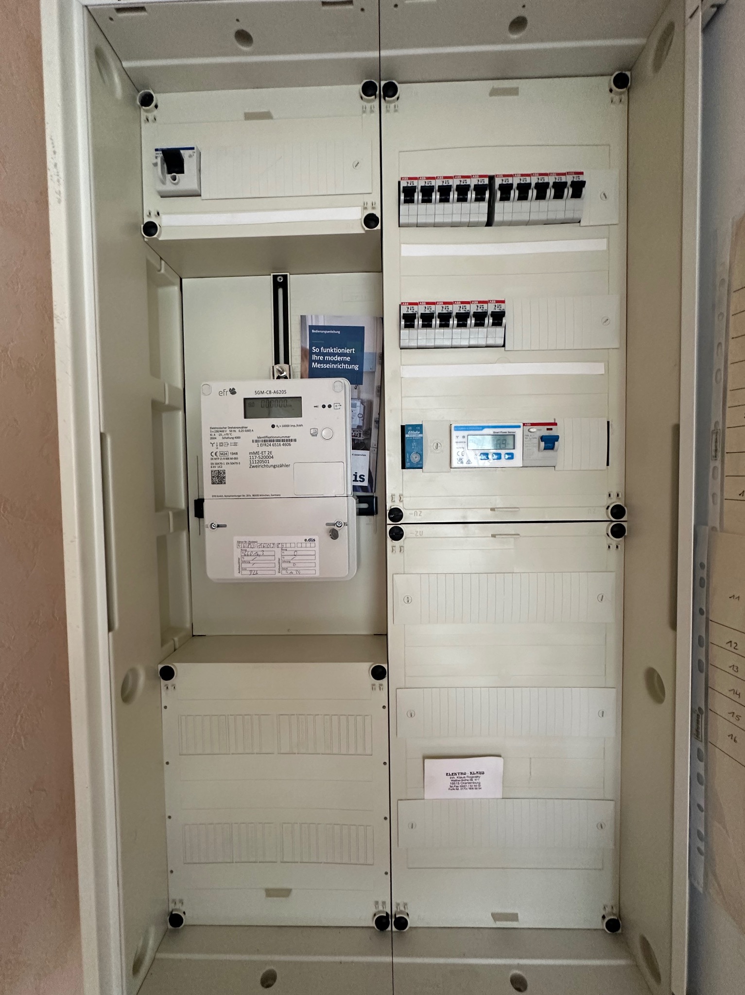 Wnętrze rozdzielnicy elektrycznej z zamontowanym licznikiem energii elektrycznej, bezpiecznikami i wyłącznikiem różnicowoprądowym. Widoczna etykieta informacyjna Elektro-Klaus.