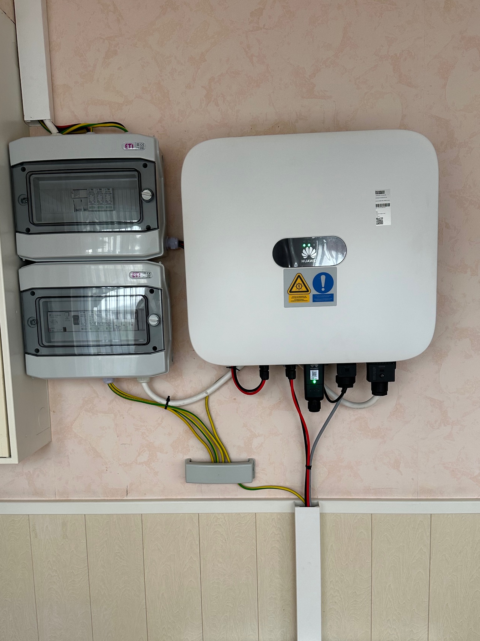 Biały inwerter fotowoltaiczny Huawei zamontowany na ścianie obok dwóch szarych rozdzielnic elektrycznych ETI, widoczne kable w kolorach: czerwonym, czarnym, żółto-zielonym, prowadzone w listwach.