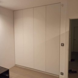 Meble Gutek - Biała, minimalistyczna szafa wnękowa z czterema frontami, zamontowana od podłogi do sufitu, z widocznymi gniazdkami elektrycznymi po prawej stronie i fragmentem ciemnego mebla po lewej.