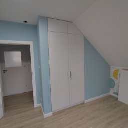 Meble Gutek - Biała, czterodrzwiowa szafa wnękowa z minimalistycznymi uchwytami, dopasowana do skosu dachu w pokoju dziecięcym z błękitnymi ścianami i drewnianą podłogą.