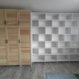 Meble Gutek - Drewniana szafa z lamelowymi drzwiami oraz biały regał z licznymi półkami w pokoju z szarą podłogą.