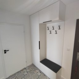 Meble Gutek - Nowoczesny przedpokój z białą zabudową meblową: szafa z czarnym uchwytem, wieszaki i siedzisko z czarną tapicerką, na podłodze wzorzyste płytki.