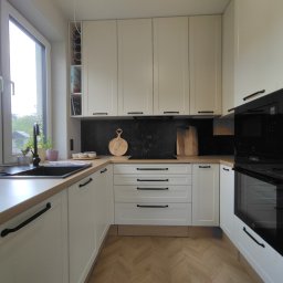 Meble Gutek - Jasna kuchnia z drewnianym blatem, czarnymi uchwytami i czarnym panelem nad blatem, z oknem z widokiem na zieleń.