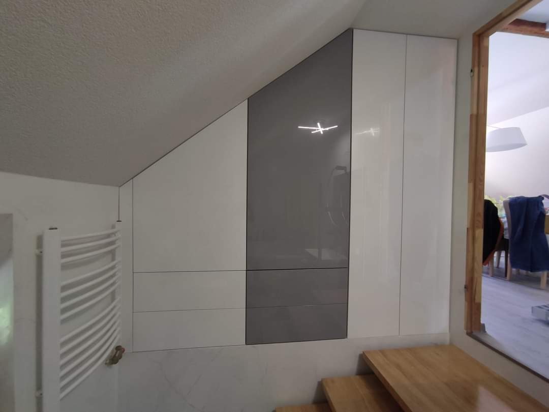 Biało-szara zabudowa meblowa na poddaszu ze ściętym sufitem, widoczny grzejnik drabinkowy i fragment drewnianych schodów.