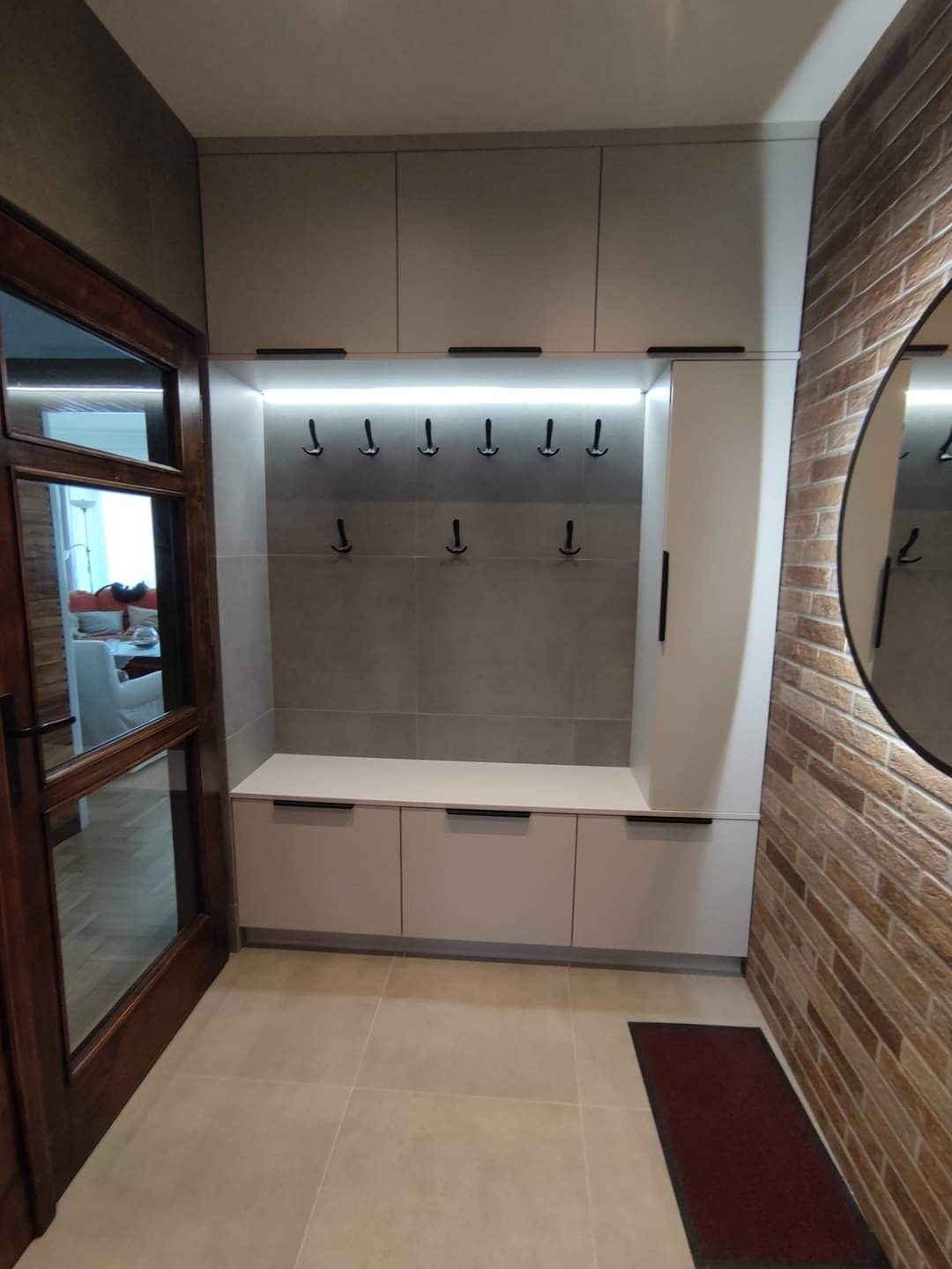 Jasnoszara zabudowa przedpokoju z wieszakami i szafkami, oświetlenie LED, ściana z cegły dekoracyjnej, widok z perspektywy wejścia.