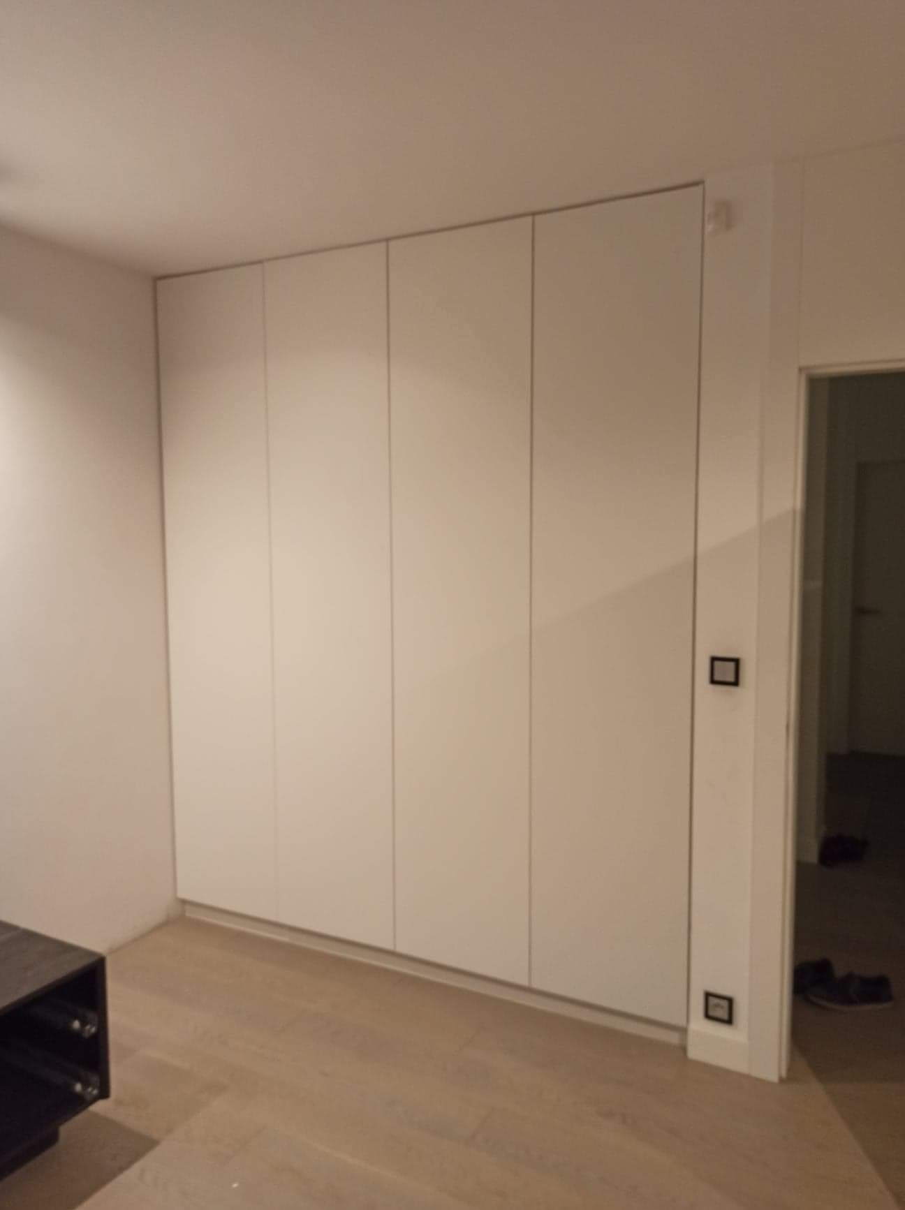 Biała, minimalistyczna szafa wnękowa z czterema frontami, zamontowana od podłogi do sufitu, z widocznymi gniazdkami elektrycznymi po prawej stronie i fragmentem ciemnego mebla po lewej.