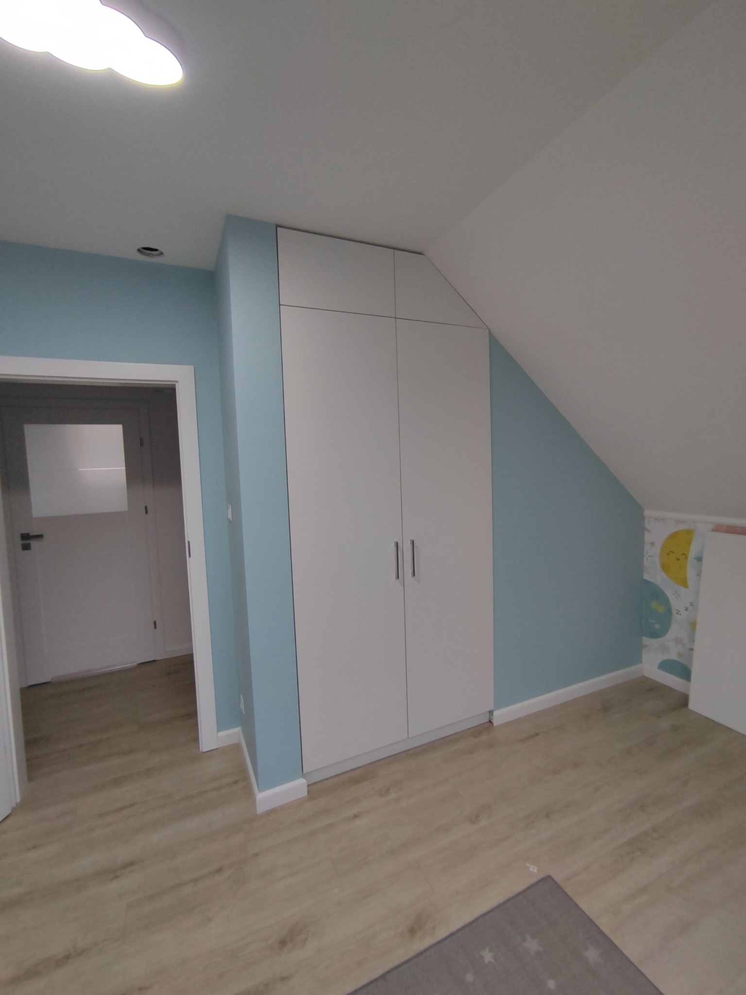 Biała, czterodrzwiowa szafa wnękowa z minimalistycznymi uchwytami, dopasowana do skosu dachu w pokoju dziecięcym z błękitnymi ścianami i drewnianą podłogą.