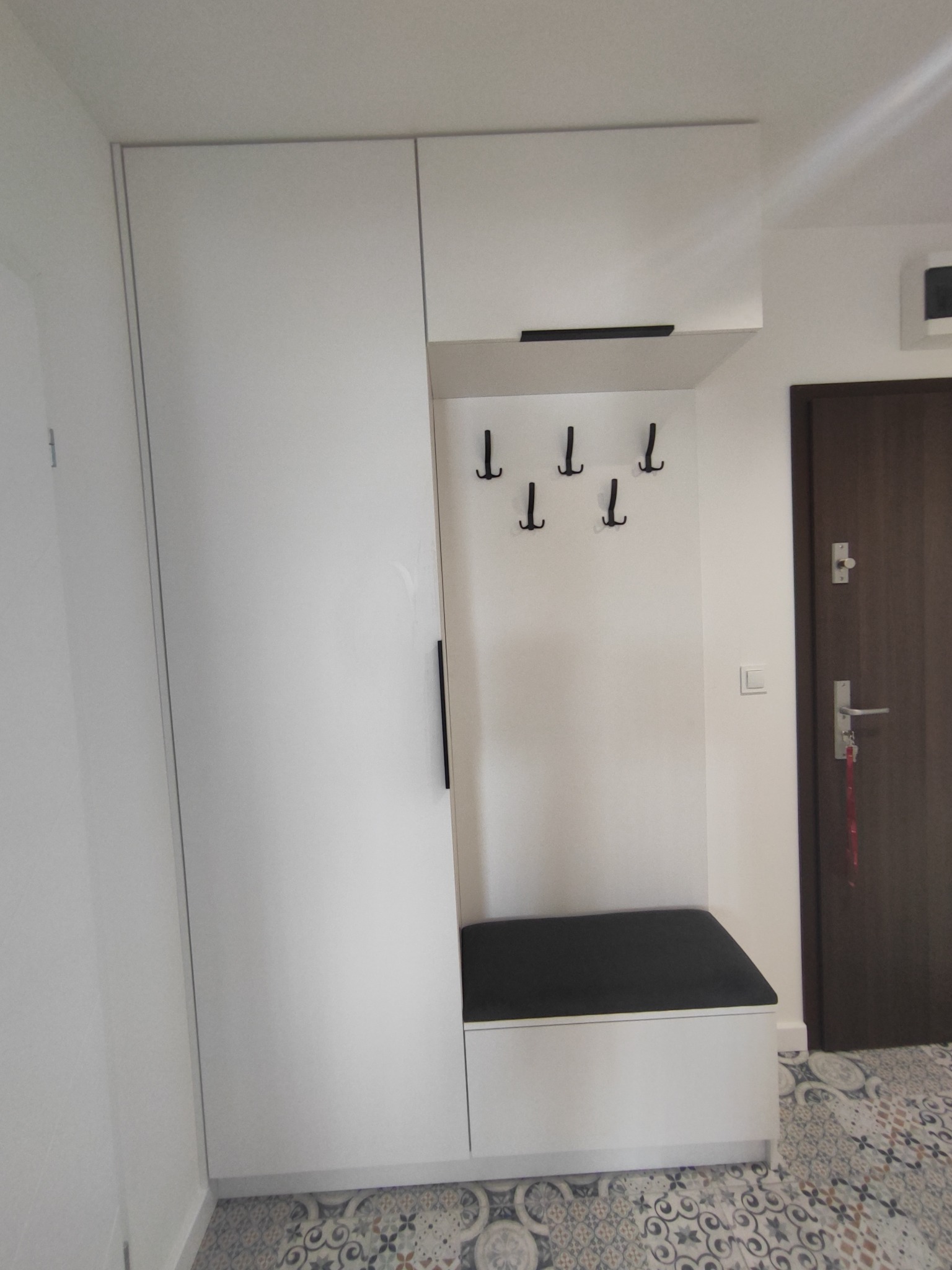 Biała zabudowa przedpokoju z siedziskiem i wieszakami w Chrzanowie, dopasowana do wzoru płytek podłogowych.