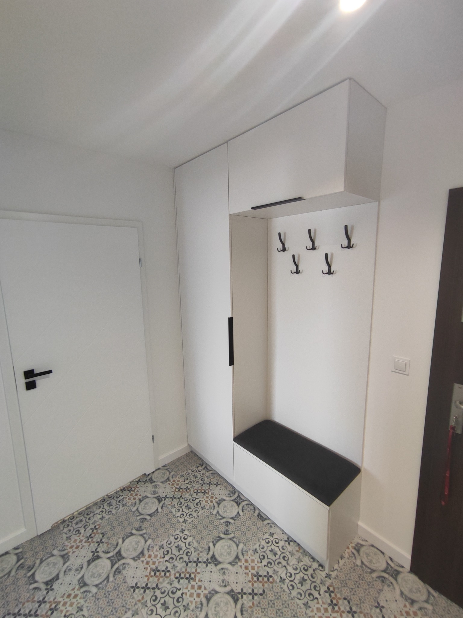 Nowoczesny przedpokój z białą zabudową meblową: szafa z czarnym uchwytem, wieszaki i siedzisko z czarną tapicerką, na podłodze wzorzyste płytki.