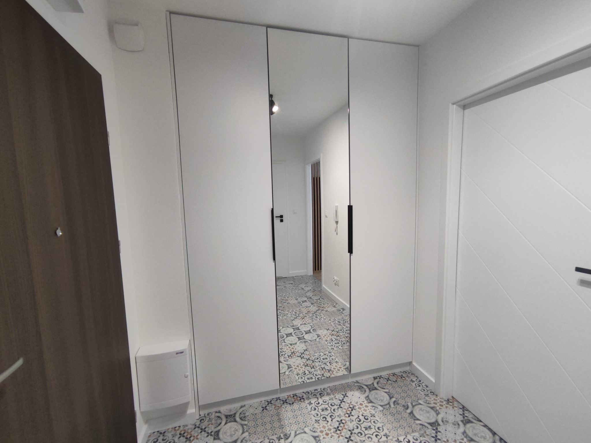 Biała szafa wnękowa z lustrzanymi drzwiami w przedpokoju z wzorzystą podłogą.