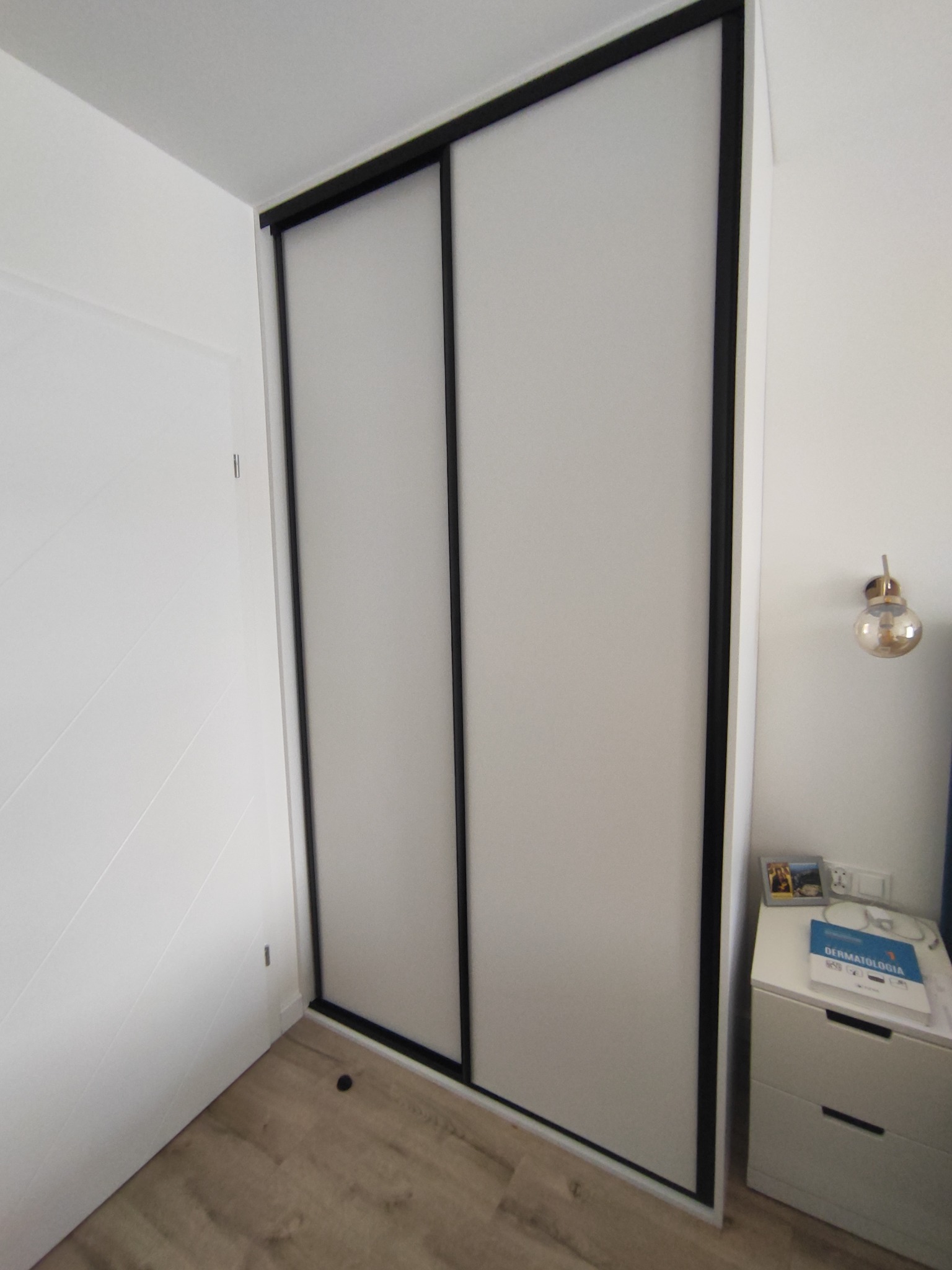 Biała szafa wnękowa z przesuwnymi drzwiami i czarnymi obramowaniami, widoczna w rogu pomieszczenia obok białej szafki nocnej z książką i lampką ścienną.