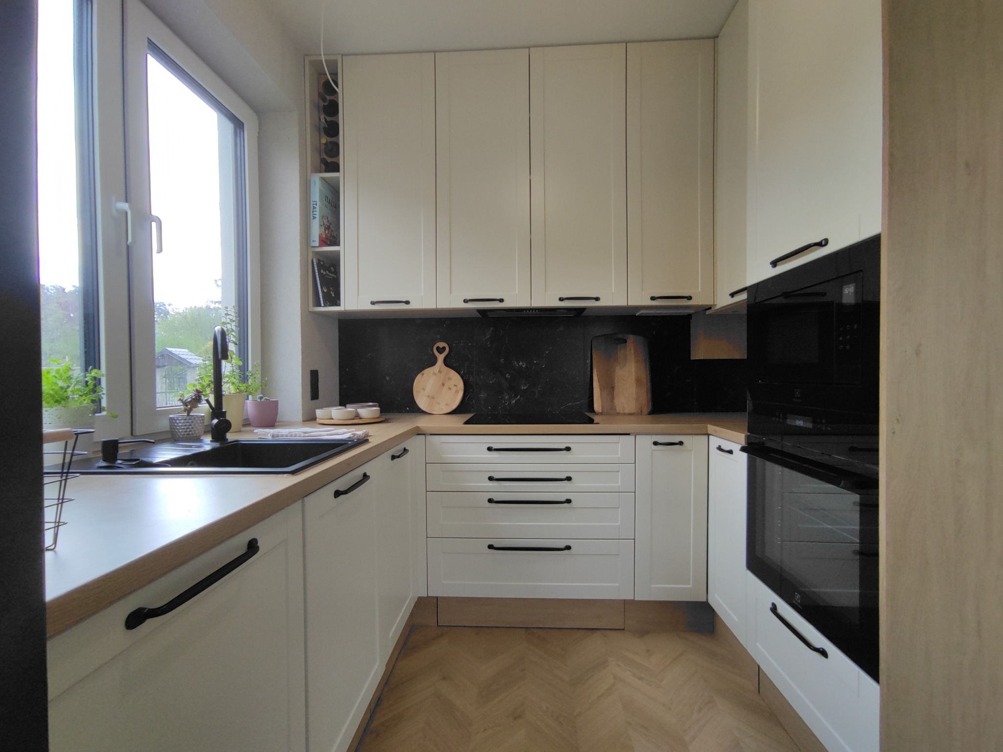 Jasna kuchnia z drewnianym blatem, czarnymi uchwytami i czarnym panelem nad blatem, z oknem z widokiem na zieleń.