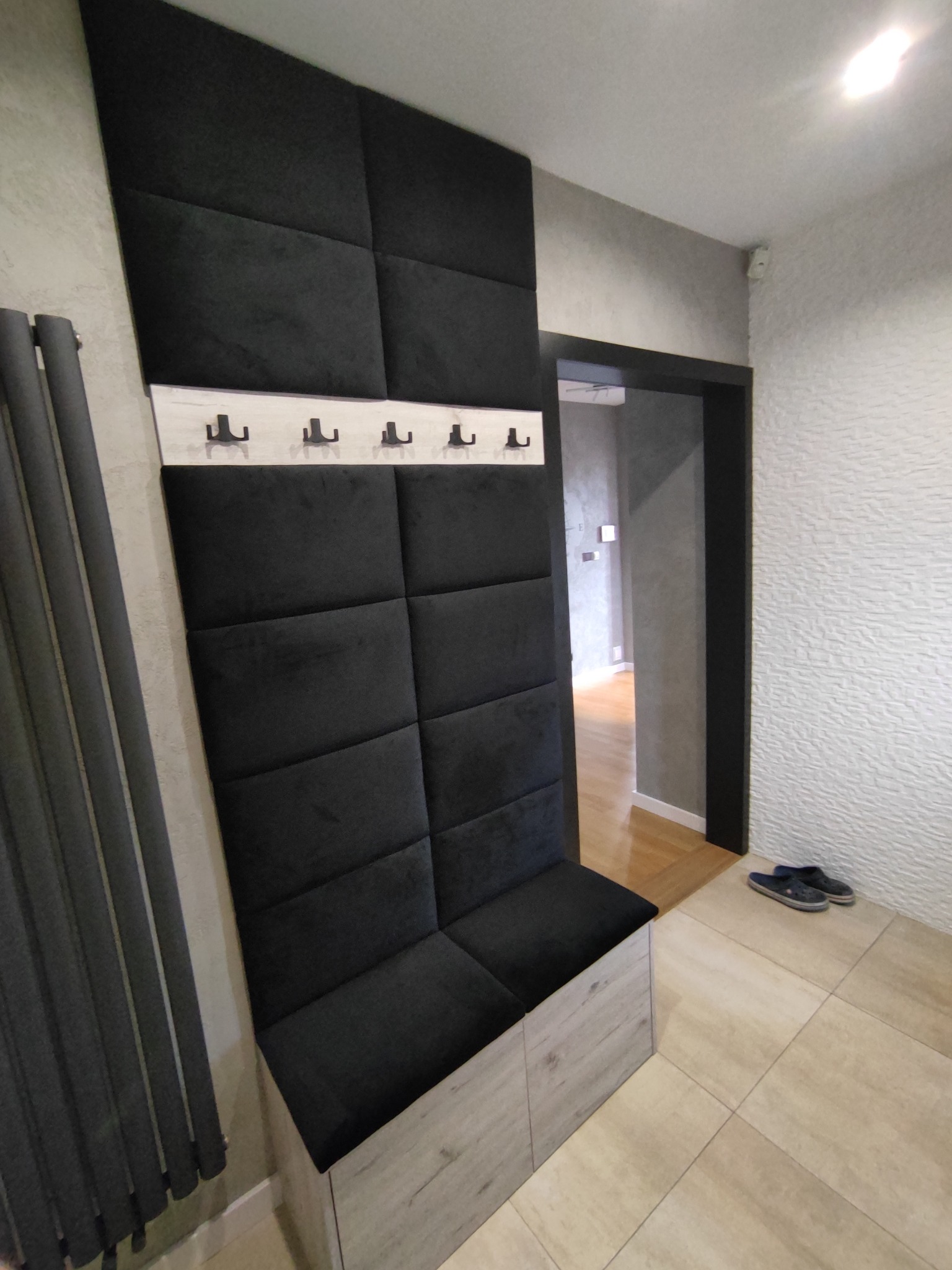 Ciemna, tapicerowana ściana z wieszakami i siedziskiem w przedpokoju, z widocznymi grzejnikiem i butami przy wejściu do innego pomieszczenia.