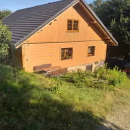 Drewniany dom z ciemnoszarą dachówką, widok z góry na ogród z ławką i stołem, słoneczny dzień.