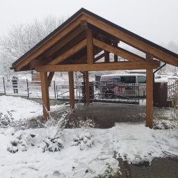 KubWood - Drewniana konstrukcja zadaszenia w zimowej scenerii, widok na bramę z kutego żelaza i częściowo zasłonięty samochód dostawczy z reklamą firmy.