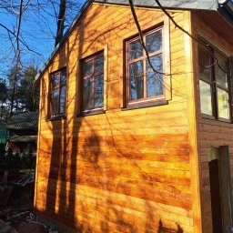 KubWood - Surowa bryła domu szkieletowego z drewnianą elewacją w trakcie budowy, widoczne okna z brązowymi ramami i fragment otynkowanej ściany, na pierwszym planie cień gałęzi.