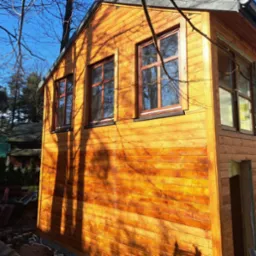 Surowa bryła domu szkieletowego z drewnianą elewacją w trakcie budowy, widoczne okna z brązowymi ramami i fragment otynkowanej ściany, na pierwszym planie cień gałęzi.