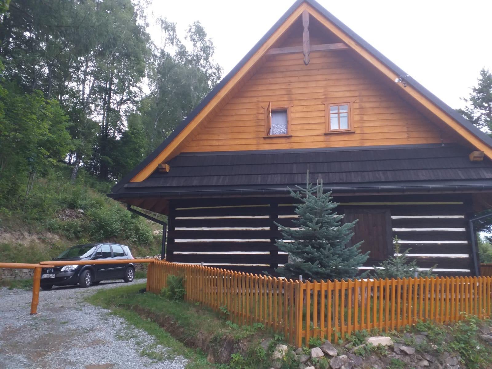 Drewniany dom w stylu góralskim z czarnym dachem i jasnymi fugami między belkami, otoczony niskim drewnianym płotkiem, z samochodem zaparkowanym obok.