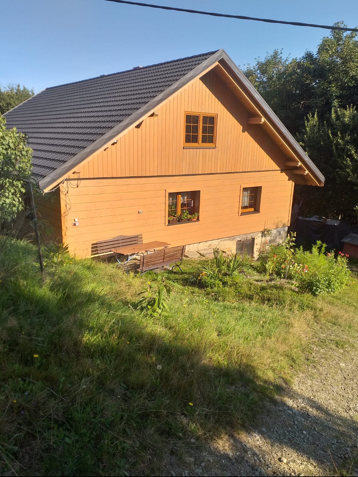 Drewniany dom z ciemnoszarą dachówką, widok z góry na ogród z ławką i stołem, słoneczny dzień.