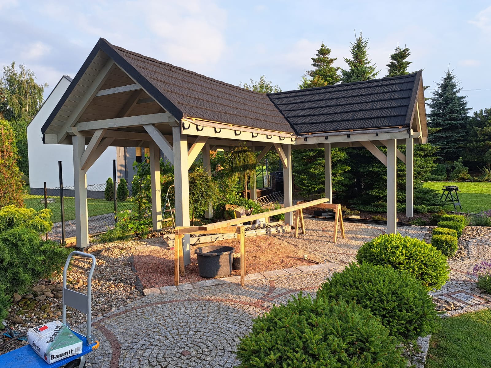 Drewniana pergola z czarnym dachem w trakcie budowy, widoczny wózek z zaprawą Baumit, narzędzia i elementy konstrukcyjne na placu budowy w ogrodzie.