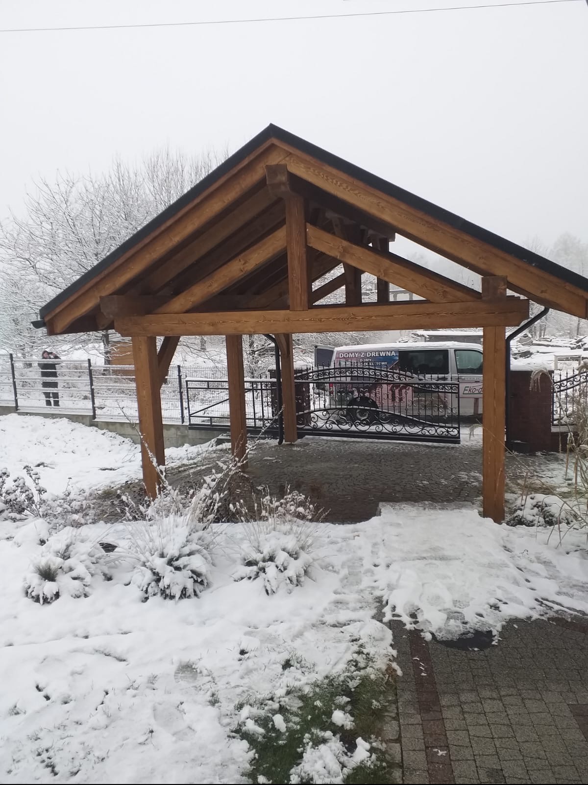 Drewniana konstrukcja zadaszenia w zimowej scenerii, widok na bramę z kutego żelaza i częściowo zasłonięty samochód dostawczy z reklamą firmy.