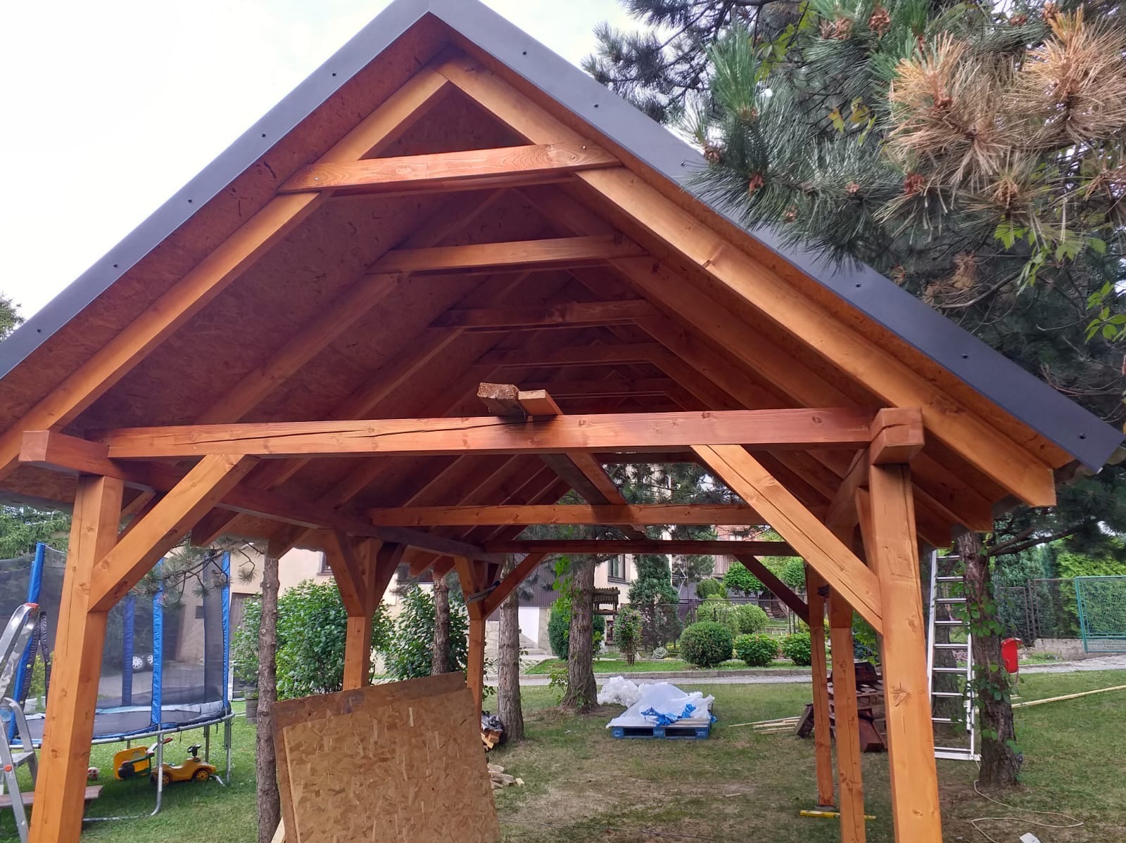 Konstrukcja drewnianej altany z widoczną więźbą dachową, czarnym obramowaniem dachu i elementami placu zabaw w tle.