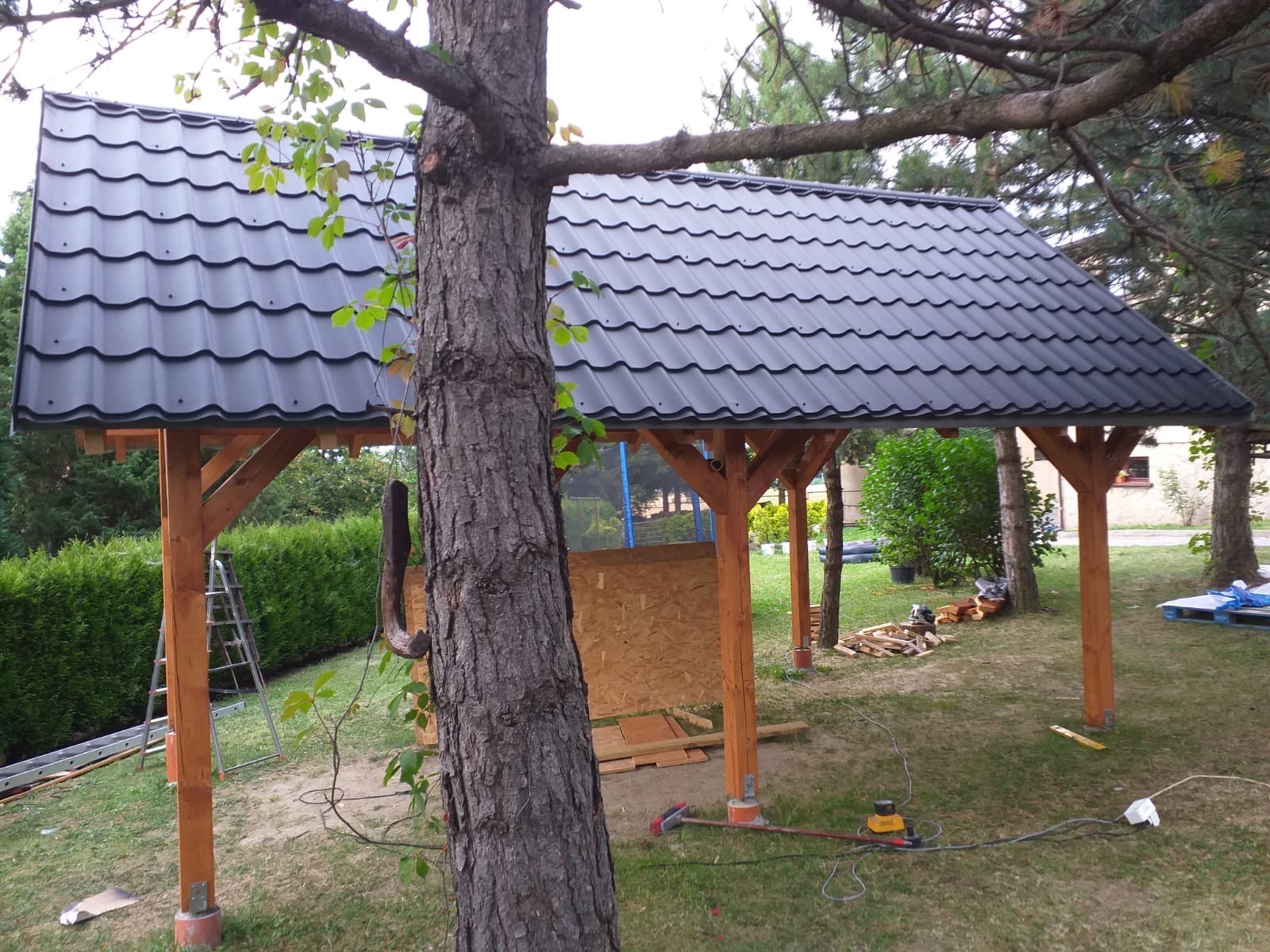 Drewniana pergola z czarnym dachem z blachodachówki w trakcie budowy w Ustroniu, widok częściowo zasłonięty przez pień drzewa, w tle narzędzia i materiały budowlane.