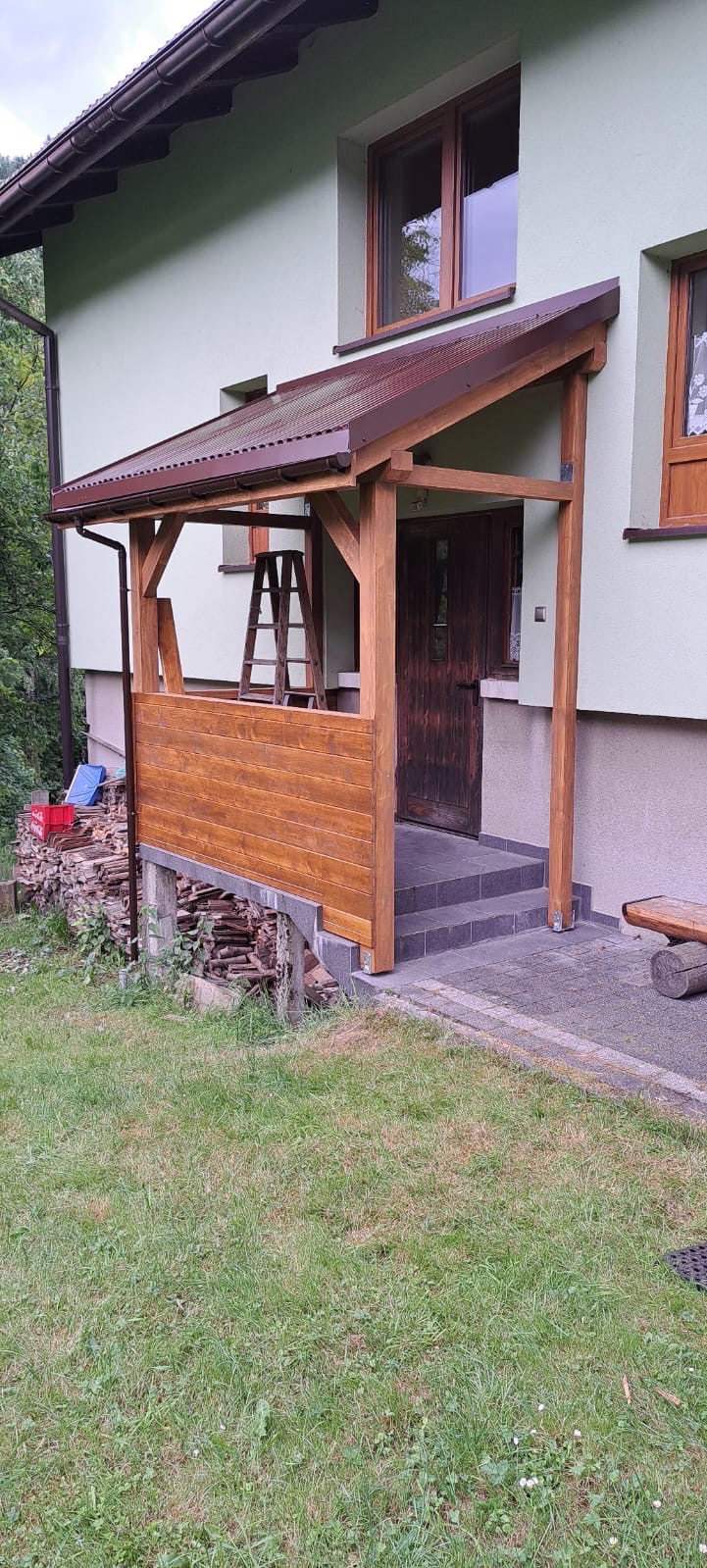 Zadaszenie drewniane nad wejściem do domu z brązowym dachem, drewnianą balustradą i schodami z szarych płytek; obok ułożone drewno na opał.