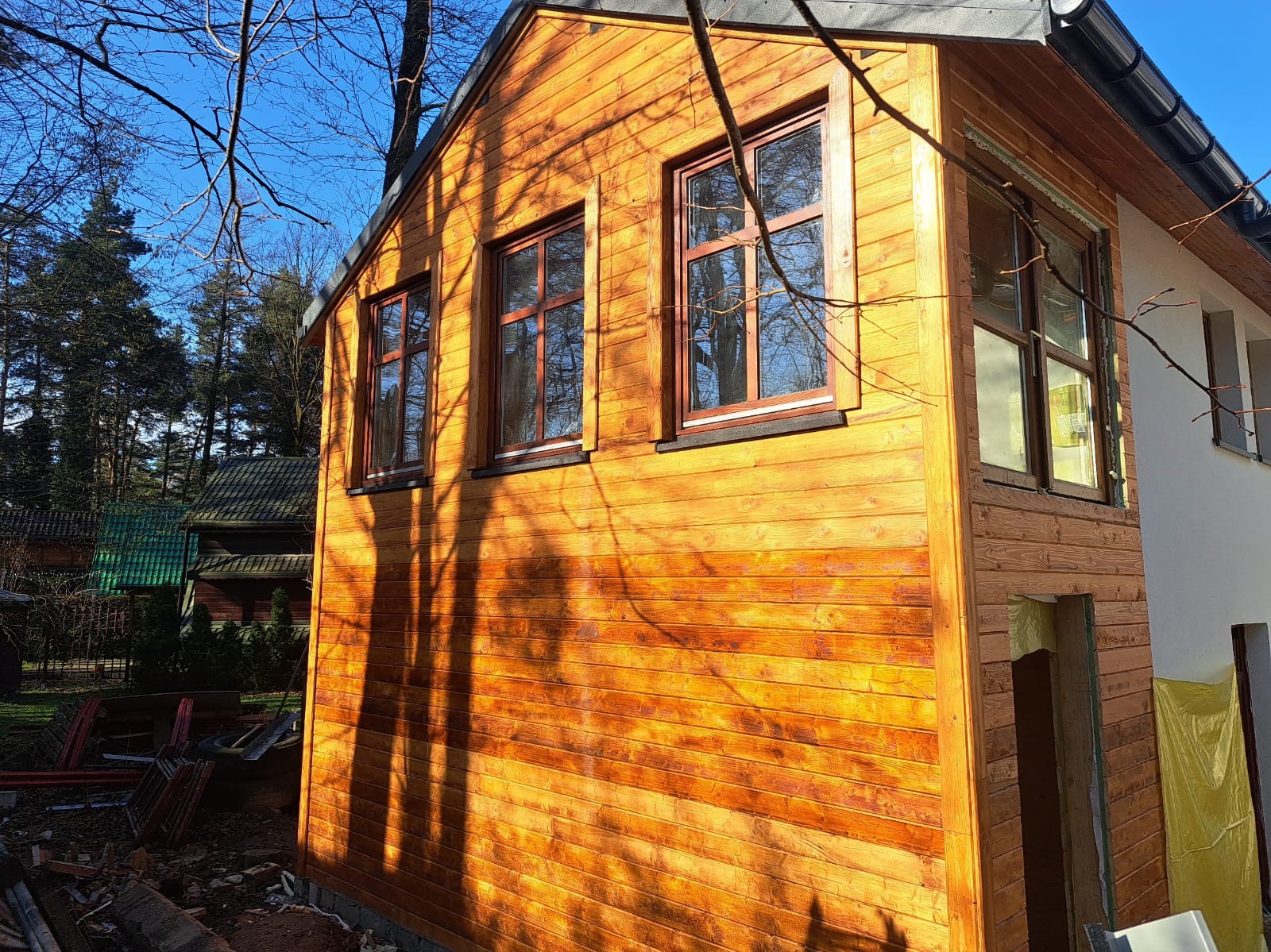 Surowa bryła domu szkieletowego z drewnianą elewacją w trakcie budowy, widoczne okna z brązowymi ramami i fragment otynkowanej ściany, na pierwszym planie cień gałęzi.