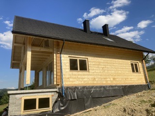 Surowy dom szkieletowy z drewnianą elewacją, czarnym dachem i rynnami, widoczny na tle górskiego krajobrazu pod błękitnym niebem.