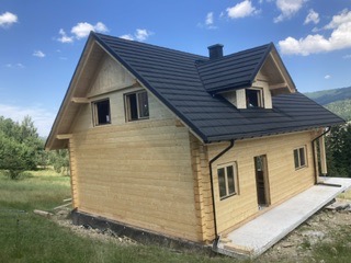 Surowy, drewniany dom szkieletowy z ciemnym dachem z blachodachówki i czarnymi rynnami, widok z boku na tle zieleni i błękitnego nieba z chmurami.